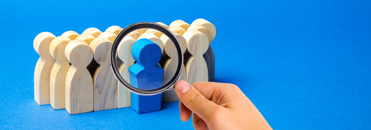 Figurines of outlines of people made from a light coloured wood are bunched together against a blue background. A magnifying glass hovers over the front figure which is blue.