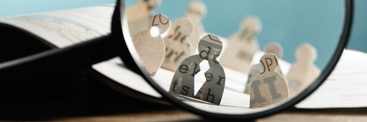 In the foreground is a mangnifying glass. Behind the magnifying glass are small outline figurines of the head and shoulder of people made from cut out pages of a book.